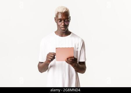 Portrait of handsome and stylish serious african-american man in glasses, looking at digital tablet, using device focused, standing white background Stock Photo