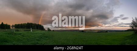 Panorama of a rainbow at sunset with red clouds over countryside with green fields, Eifel, Germany Stock Photo