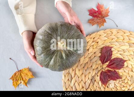 Female hands holding a beautiful pumpkin on gray surface with dry autumn leaves and a wicker napkin Stock Photo