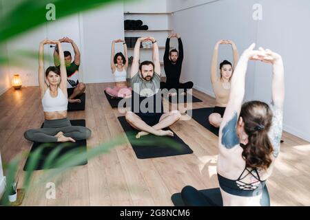 Hard working people practicing yoga in a group, doing stretches in easy pose in a big studio. In front of an instrustor, girl with shoulder tattoo. Stock Photo