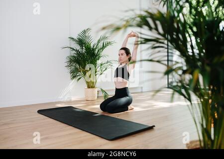 Serious, focused woman in black sportswear practicing yoga, sitting on her legs, stretching in a big studio. Side view Stock Photo