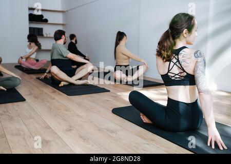 People practicing yoga in a group, twisting in easy pose, stretching in a big studio. In front of an instrustor, girl with shoulder tattoo. Stock Photo