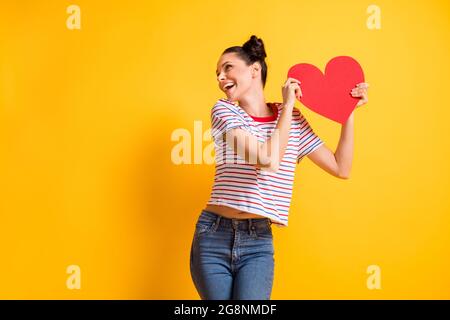 Photo of sweet cute young lady wear striped t-shirt dancing holding big red heart looking empty space isolated yellow color background Stock Photo