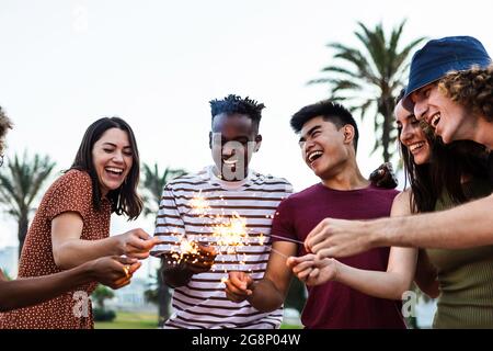 Happy multiracial people enjoying summer party at the beach - Diverse group of young adult students celebrating together with sparklers outdoors - Stock Photo