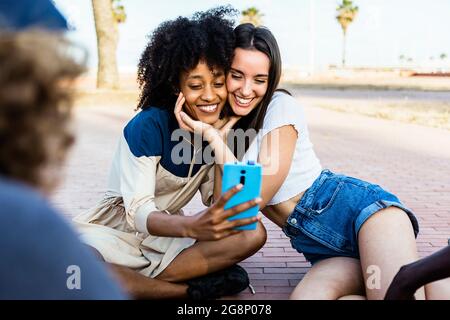 Happy multiracial women friends taking a selfie with smart phone outdoors - Joyful multiethnic female couple having fun with their friends Stock Photo