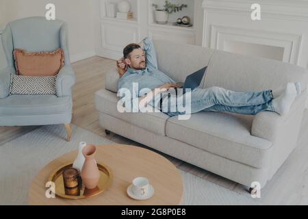 Young businessman working remotely at home while lying on couch Stock Photo