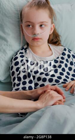 Hospitalized child wearing oxygen nasal tube resting in bed with medical oximeter on finger measuring heartbeat pulse. Worried mother discussing with kid during therapy consultation in hospital ward Stock Photo