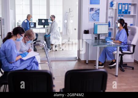 Nurse checking appointment form of old disabled woman with walking frame sitting in waiting room wearing protection masks against coronavirus. Patients visiting doctor during covid 19 global pandemic Stock Photo
