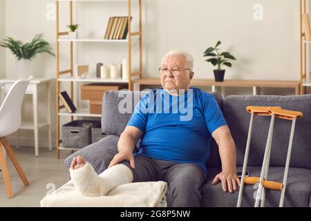 Disappointed senior mature man sitting on sofa with broken leg in cast Stock Photo