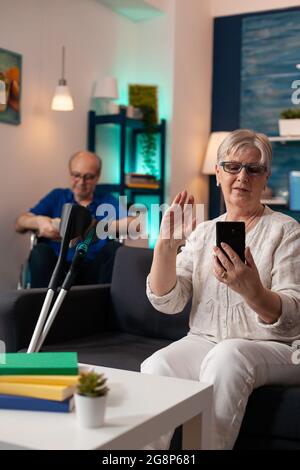 Mature retired couple on modern video call at home using smartphone technology online conference. Elder man reading while senior woman talking to family on webcam internet connection Stock Photo