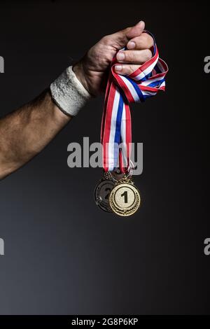 Arm of elderly athlete holding three gold, silver and bronze medals on a dark background. Sports and victory concept. Stock Photo