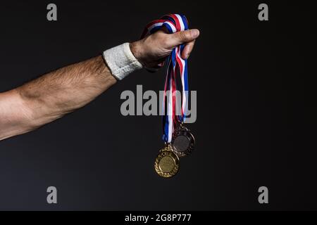 Arm of elderly athlete holding three gold, silver and bronze medals on a dark background. Sports and victory concept. Stock Photo