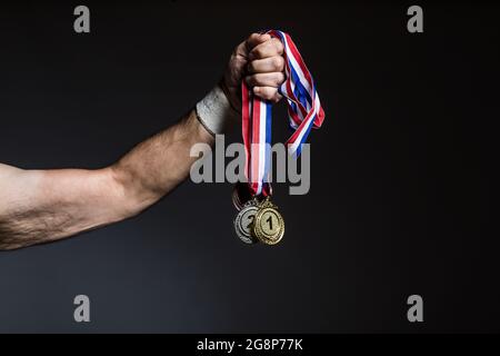 Arm of elderly athlete holding three gold, silver and bronze medals on a dark background. Sports and victory concept. Stock Photo