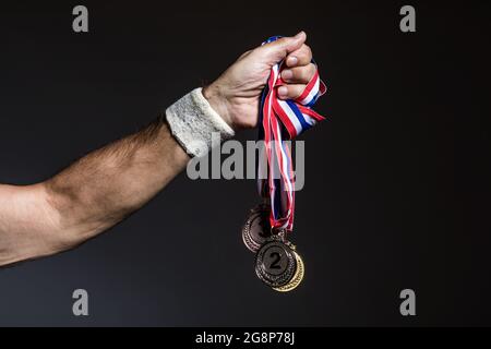 Arm of elderly athlete holding three gold, silver and bronze medals on a dark background. Sports and victory concept. Stock Photo