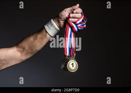 Arm of elderly athlete holding three gold, silver and bronze medals on a dark background. Sports and victory concept. Stock Photo