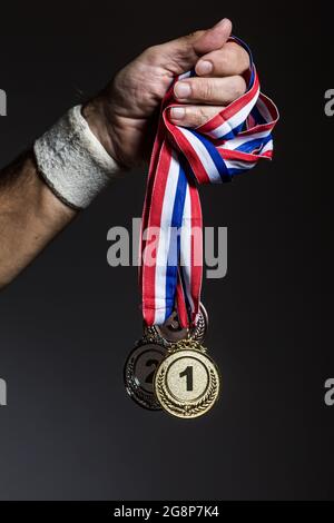 Arm of elderly athlete holding three gold, silver and bronze medals on a dark background. Sports and victory concept. Stock Photo