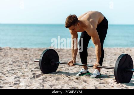 Muscular guy bodybuilder doing exercises with big dumbbells outdoors on the beach, on a summer sunny day, training on the sea coast on the background Stock Photo