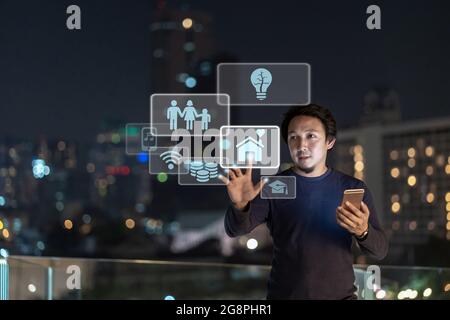 Asian creative freelancer using smart mobile phone with smile action touching the virtual reality of House and basic factor icon over the cityscape ba Stock Photo
