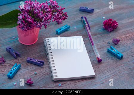 Blank notebook, pencils, lilac flowers and colorful pegs on vintage wooden painted blue background. Stock Photo
