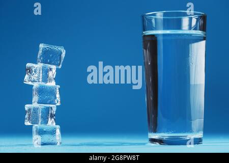 A glass of refreshing water and a tower of ice cubes on a blue background. A refreshing drink on hot days. Free space Stock Photo