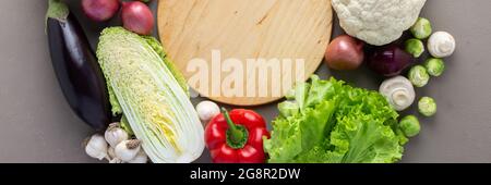 Fresh farm organic vegetables banner, healthy food concept, vegetables and mushrooms around a round cutting board on a gray background, top view Stock Photo