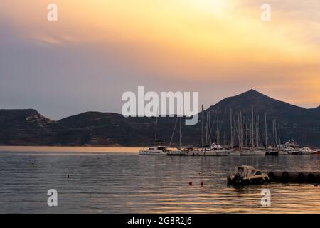 Fishing and sailing boats docked in Adamas Port on Milos Island, Greece during golden hour sunset, with calm flat sea and orange cloudy sky. Stock Photo