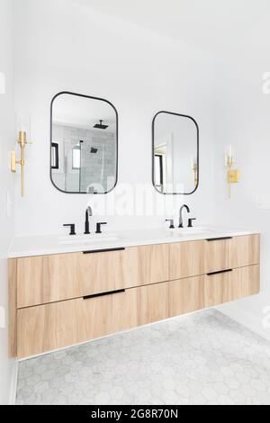 A beautifully renovated bathroom with a wood cabinet, black framed mirror and faucets, and gold lights mounted on the wall. Stock Photo