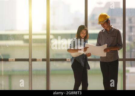Two Asian engineer, male with yellow safety helmet and woman with white one standing and talking near high curtain wall glass frame in construction si Stock Photo