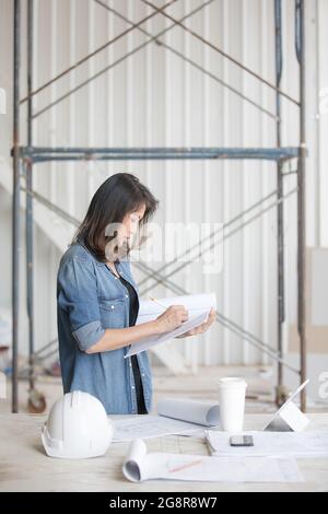 Beautiful Asian engineer woman in blue jeans shirt standing and writing, white safty helmet on desk. Stock Photo