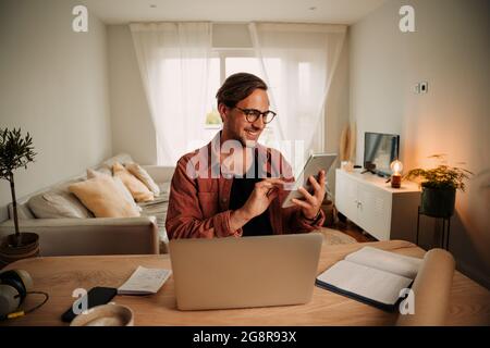 Caucasian male student typing on digital tablet smiling working from home wearing spectacles Stock Photo