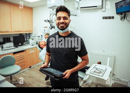 Mixed race male nurse standing holding digital tablet standing in doctors office with patient and female dentist holding digital tablet Stock Photo