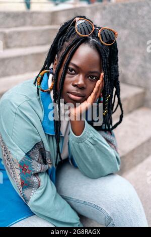 Expressive young african woman looking at camera outdoors and posing. She is sitting in stairs in the city. Stock Photo
