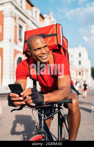 Smiley messenger man sitting in his bike using his phone in his way to deliver a package. Stock Photo