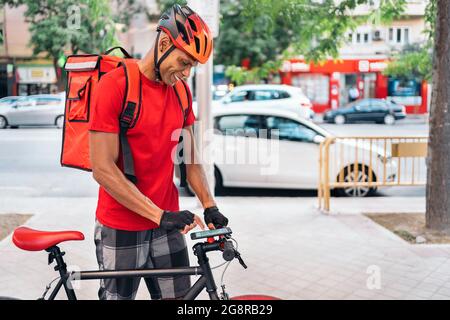 Happy african messenger man using his phone in the street in his way to deliver a package. Stock Photo