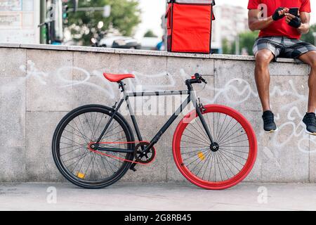 Unrecognized delivery man sitting in the street next to his bike and using his mobile phone. Stock Photo
