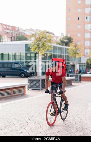 Strong african american delivery man riding his bike and carrying a package in the city. Stock Photo