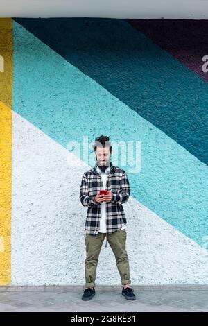 Happy hipster man standing against colorful wall in the city and using his phone. Stock Photo