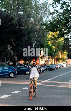 Happy hipster man with big beard riding his bike in the city and raising his arms. Stock Photo