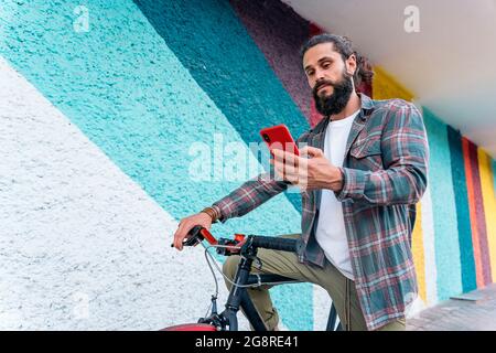 Young hipster man with long hair smiling and using his smart-phone while sitting in his bike. Stock Photo