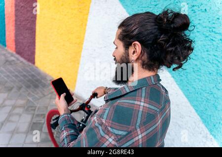 Cool hipster man with long hair smiling and using his smart-phone while holding his bike. Stock Photo