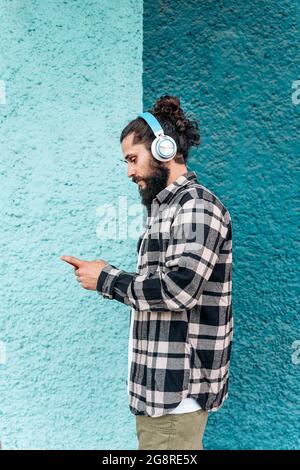 Confident hipster man standing against blue wall in the city and using his phone. He is wearing headphones. Stock Photo
