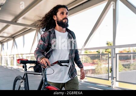 Cool bearded man with long hair smiling and looking at front while holding his bike. Stock Photo