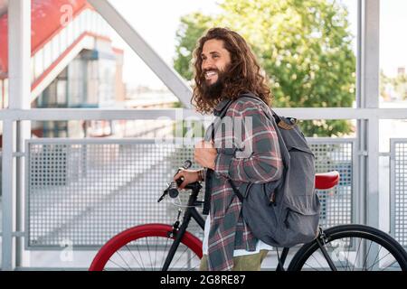Happy man with long hair walking in the city with his bike and looking at front. Stock Photo