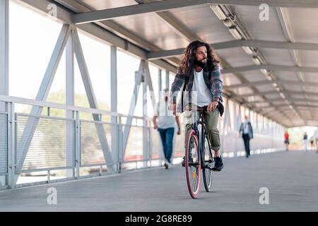 Handsome hipster man with long hair looking to the side and riding his bike in the city. Stock Photo