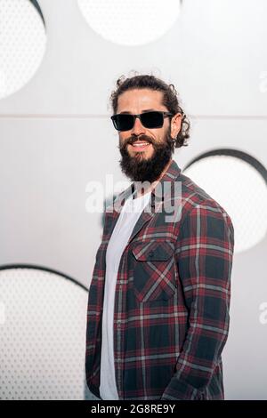 Confident hipster man wearing sunglasses smiling and looking at camera against white background. Stock Photo