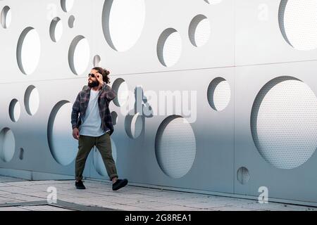 Young man with long beard and wearing sunglasses walking in the street against white wall with cool design. Stock Photo