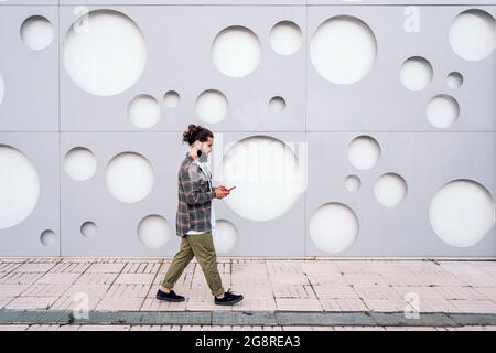 Young man with long beard walking in the street against white wall with cool design and using his phone. Stock Photo
