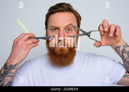 Worried man with scissors and blade is ready to cut the beard Stock Photo