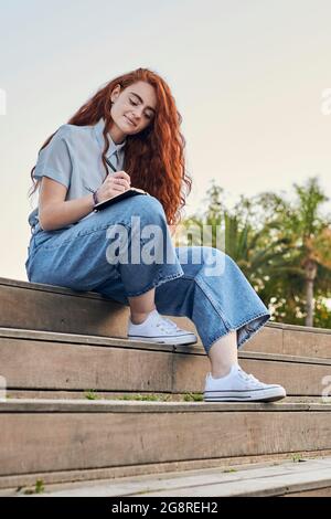 Young redhead woman writes in a notebook outdoors Stock Photo