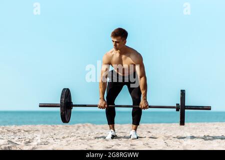 Healthy handsome active man with fit muscular body, young muscular man lifting weights, doing exercises, Sporty man exercising on the beach, Outdoor t Stock Photo